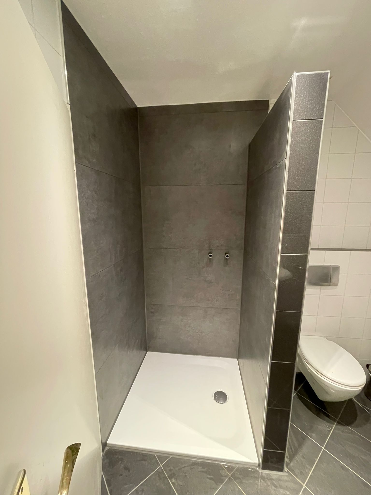 Bodengleiche Dusche in Betonoptik mit weisser Duschwanne und dunklem Mosaikstreifen neben dem Wand-WC